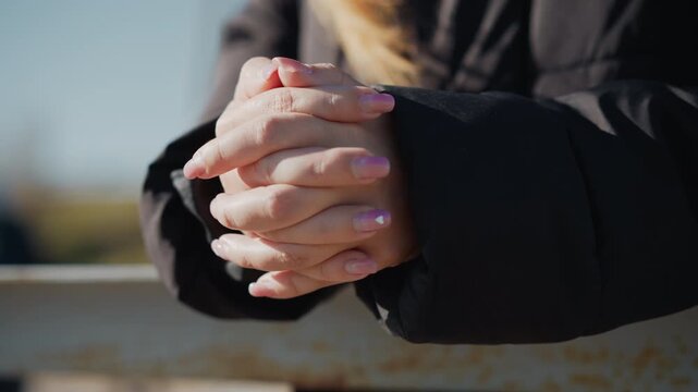 Woman with pink heart nail design clasps hands together in close up wearing black jacket outdoors under bright sunlight. Hands remain centered showing consistent hand positioning