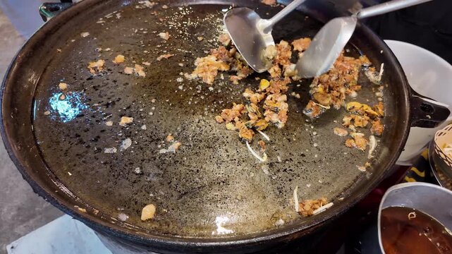 Close up shot of vendor hands using two spatulas to fry traditional crispy Hoi Tod mussel pancake on a flat pan at Thailand market.