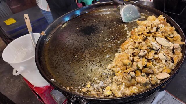 Close up shot of freshly cooked crispy Hoi Tod or Thai mussel pancake with seafood on a large flat griddle at a market stall.
