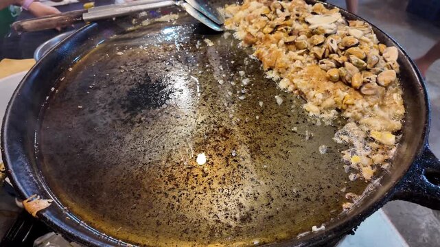Close up shot of freshly fried traditional Hoi Tod mussel pancake resting on a flat iron pan at a Thailand street food market.
