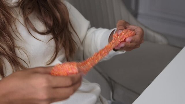 Close-up of young person's hands stretching and playing with textured orange slime with small beads, indoors.