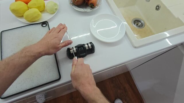 Point of view shot of a person sharpening a metal kitchen knife using a manual sharpener on a white countertop surrounded by fresh vegetables before cooking a meal at home.