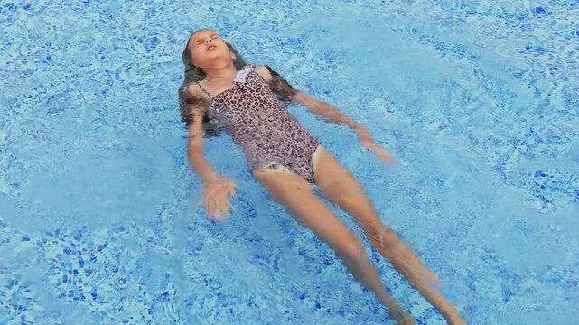 Young girl in a leopard print swimsuit floating serenely on her back with eyes closed in clear blue swimming pool water.