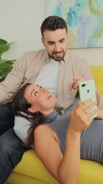 A couple lounges on a yellow sofa, taking a buoyant selfie in a sunlit living room. One partner leans back with a relaxed smile while the other holds the phone, capturing a candid, cheerful moment.