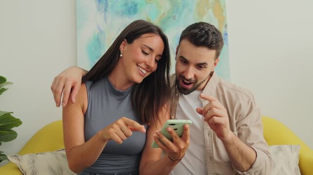 A loving couple sits close on a sofa, engaging with a smartphone. They laugh and point, showing collaboration and enjoyment in a cozy living space with soft light and pastel decor.