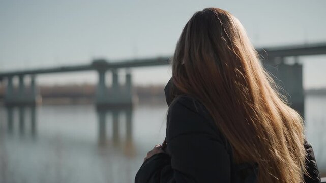 Woman with long hair stands by body of water wearing dark outerwear and carrying item in her hands. background features soft daylight reflections on calm surface below and distant structures