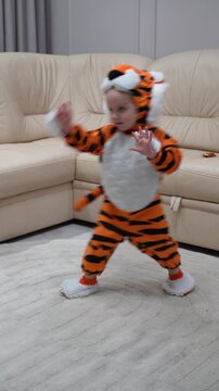 Young child dressed in tiger costume playfully dances and moves around in living room setting with large sofa and rug.
