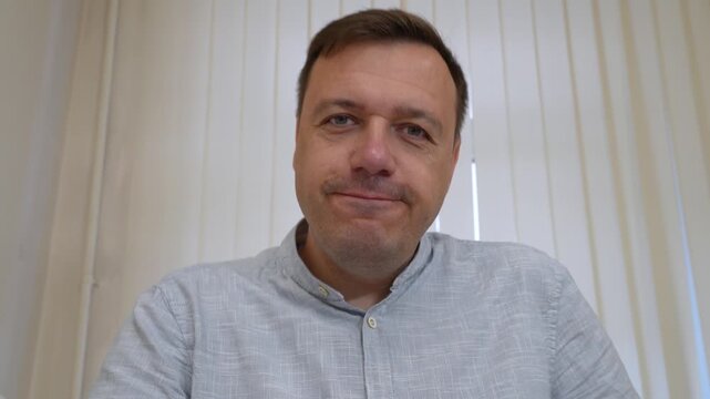 Close-up of middle-aged man in light blue collared shirt looking directly at camera and speaking, with vertical blinds in background.