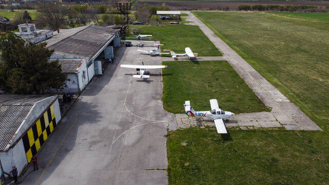 Serbia - 11 April 2021: Aerial view of light aircraft parked on the concrete apron of a small airfield with a hangar and surrounding green fields.