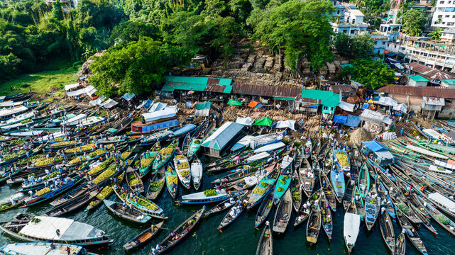 Rangamati, Bangladesh - 19 October 2022: Aerial view of the bustling boat market with numerous wooden boats laden with goods docked along the shore, surrounded by lush trees.