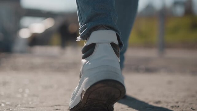 Woman walks steadily on paved path wearing white sneakers and blue jeans. focus remains consistently low angle at her feet throughout sequence capturing subtle movements of each step