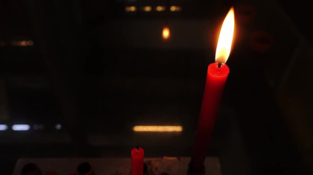 A bright red candle flame illuminates a dark scene, casting reflections on a smooth, dark surface with subtle background light sources, creating a mysterious and contemplative atmosphere