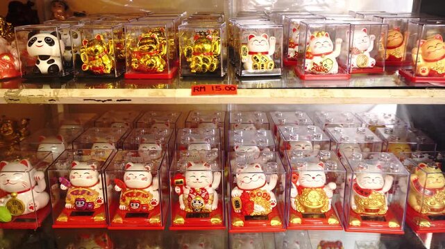 Collection of assorted waving cat figurines and a panda figurine in clear plastic boxes on shelves, with a price tag visible