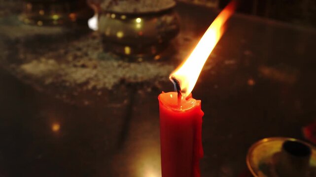 A red candle burns with a bright flame, casting a warm glow on a dark, textured surface with a golden incense burner in the background