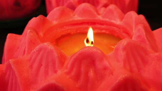 A close-up shot of a vibrant red-orange candle with a flame burning in the center, casting a warm glow on the textured wax petals against a dark background