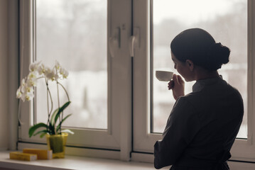 Fototapeta premium Silhouetted woman in dark uniform stands near window, holding small cup close while looking outside, soft natural backlight outlining profile, bun, and quiet interior mood with blurred winter view