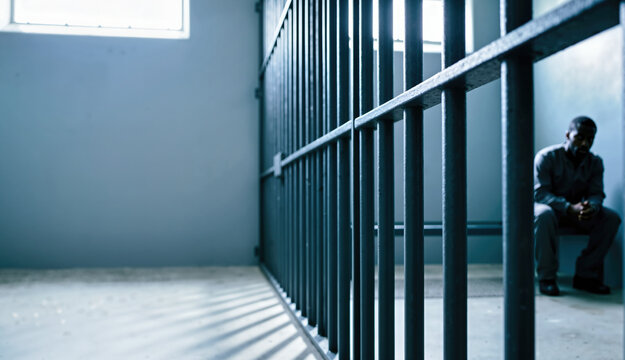 Solitary prisoner sitting behind bars in a dark cell. Black male inmate in gray uniform in a muted blue prison interior