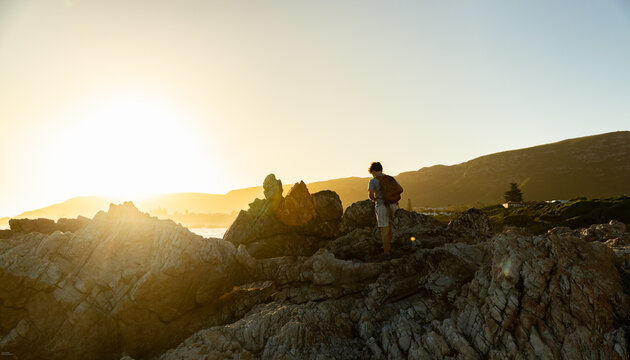 13 year old boy wearing backpack exploring the iconic rocky coastline of Kammabaai Beach, Hermanus, South Africa at sunset