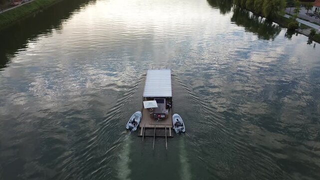 Aerial drone view of a small river boat cruising along the Drava River in Maribor, Slovenia, with calm reflective water, riverside greenery and warm evening sunlight.
