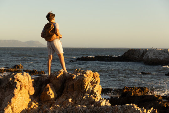 13 year old boy wearing backpack exploring the iconic rocky coastline of Kammabaai Beach, Hermanus, South Africa at sunset