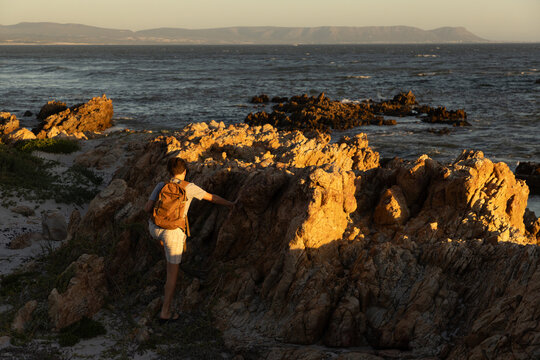 13 year old boy wearing backpack exploring the iconic rocky coastline of Kammabaai Beach, Hermanus, South Africa at sunset