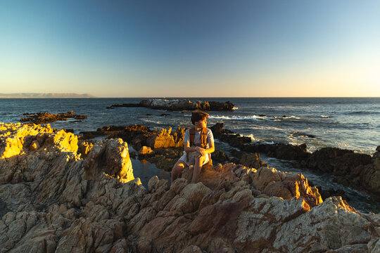 13 year old boy wearing backpack exploring the iconic rocky coastline of Kammabaai Beach, Hermanus, South Africa at sunset