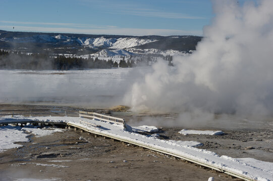 View of a geyser erupting with steam in a snow-covered landscape with a wooden boardwalk in the foreground at Yellowstone National Park, Wyoming, United States.