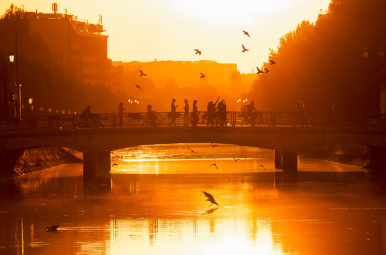 View of people walking and cycling across a bridge over the Dambovita River during a golden sunset in Bucharest, Bucharest, Romania.