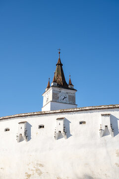 View of the historic clock tower of the Prejmer Fortified Church rising above white defensive walls against a clear blue sky in Prejmer, Brasov, Romania.