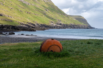 A rusty orange buoy rests in the grass beside a black sand beach and green cliffs in the Faroe Islands. Maritime heritage and wild coast for travel and nautical themes. © Oksana