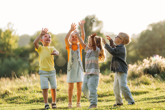 Four children reaching upward together in a sunlit meadow while playing outdoors, joyful summer scene with bubbles and grass, candid childhood friendship portrait