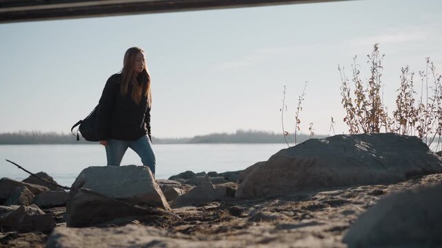 Young woman walks along rocky riverside path under overpass carrying black backpack. Woman wears casual clothing and appears to be exploring outdoor environment with water and distant