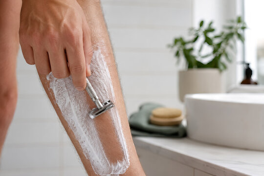 A person shaves his legs with a straight razor in a bright, modern bathroom. The human is positioned in front of a white sink with a circular vessel and a pale grey countertop.