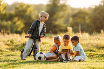 Obraz premium Children gathering on grass with scooter, soccer ball, and tablet during outdoor play in a sunlit park, candid childhood scene with trees and meadow background