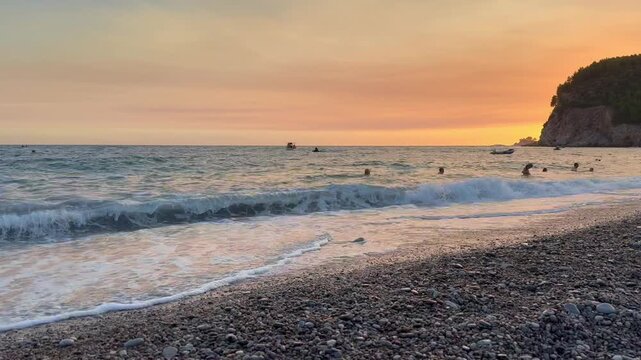 A beautiful view on Adriatic sea and Buljarica beach on sunset, Montenegro. Gentle waves roll onto a pebbled beach as swimmers move through the sea beneath a glowing sunset beside a rocky coastal
