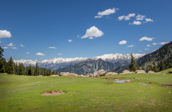 View of the meadow landscape near Jahaz Banda Alpine Rest House with snow-capped mountains in the background under a clear blue sky in Kumrat Valley, Khyber Pakhtunkhwa, Pakistan.