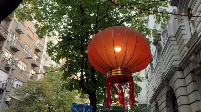 Glowing red Chinese lantern hanging above an urban street with tassels, trees, historic buildings, and overhead decorations in a city neighborhood. Chinese New Year decor.