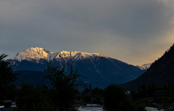 View of snow-capped mountains of Kumrat Valley illuminated by golden hour light under a dramatic cloudy sky in Kumrat Valley, Khyber Pakhtunkhwa, Pakistan.