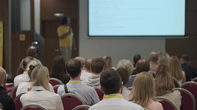 Business conference audience listening attentively to presenter, projected slides visible, networking and learning atmosphere.