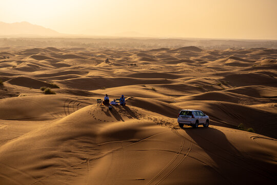 View of Erg Chebbi sand dunes with two people sitting on chairs and a white SUV parked in the desert landscape during golden hour in Errachidia Province, Draa-Tafilalet, Morocco.