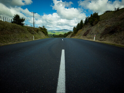 View of an asphalt road stretching through the rolling green hills under a bright blue sky with scattered clouds in Waikino, Waikato Region, New Zealand.