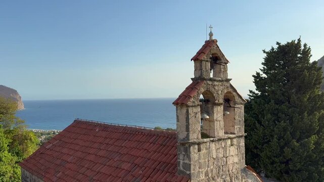 A beautiful view on Gradiste Monastery Church bell tower, Orthodox complex, Buljarica, Montenegro. Stone church bell tower with red tiled roof overlooking blue sea and coastal mountains in peaceful