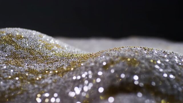 Shimmering gold and silver mesh fabric forms soft rolling waves as reflective particles sparkle across the textured surface against a deep black background with shallow focus