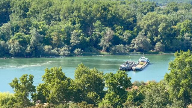 Cargo barges moving along a wide river bordered by dense green woodland and trees, viewed from an elevated distance in bright summer daylight, peaceful inland waterway landscape
