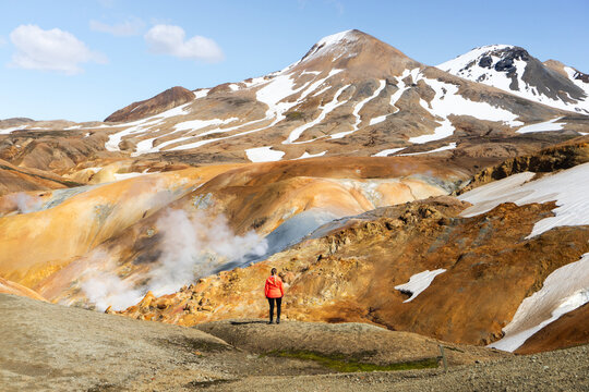 View of a hiker in an orange jacket standing in the geothermal landscape of Kerlingarfjoll with steaming vents, colorful rhyolite mountains, and patches of snow in Hrunamannahreppur, Iceland.