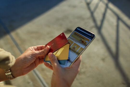 Woman's hands with a watch hold a smartphone with an online store website open and several credit cards (red, blue, gold) outdoors. Selecting a card for a payment.