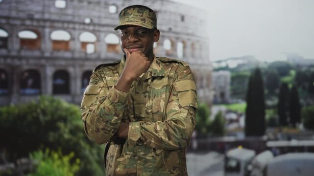 Man in camouflage military uniform and cap touches chin with hand while standing before a historic building; pride duty.