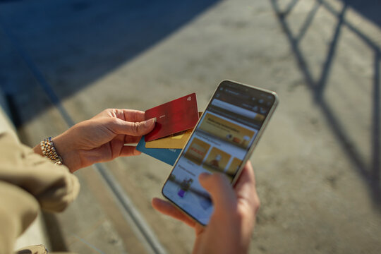 Close-up of a woman's hands holding a smartphone with an online store website open and several credit cards outdoors. Selecting a bank card for a secure mobile payment in bright sunlight.