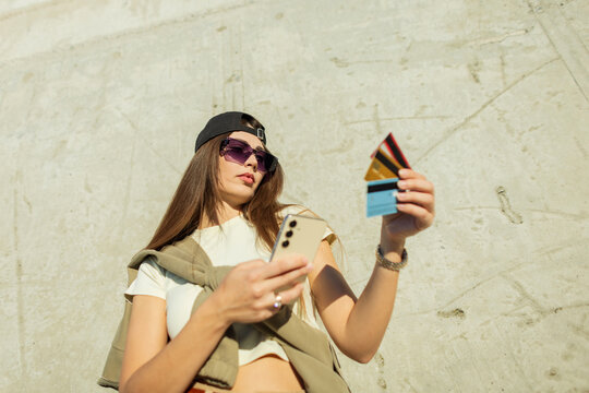 young stylish woman makes an online purchase using her smartphone and multiple credit cards, shot from a dynamic low angle against a textured urban concrete wall in bright sunlight. E-commerce