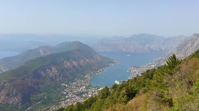 A beautiful view on Kotor Bay, Montenegro. Aerial panoramic view moves across rugged coastal mountains, blue bay, forested hills, islands, and clustered seaside settlements under hazy summer skies in